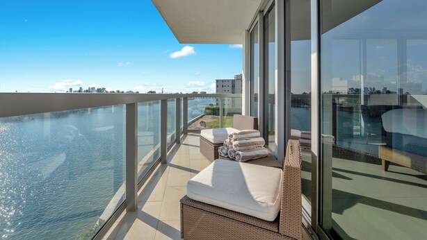 A balcony with wicker furniture, a glass railing, and a view of the water and city skyline. - Stunning 2BR 2BA  Bay Harbour with Pool (Bay Harbor Islands)