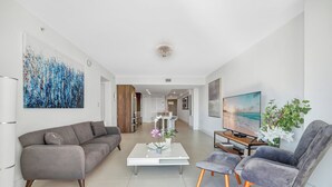 A modern living room with a grey sofa, a white coffee table, and a grey armchair. There is a TV on a wooden stand and a large abstract painting on the wall. - Stunning 2BR 2BA  Bay Harbour with Pool (Bay Harbor Islands)
