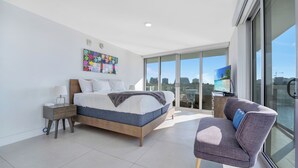 A modern bedroom with a large bed, a nightstand, a chair, and a city view through large windows. - Stunning 2BR 2BA  Bay Harbour with Pool (Bay Harbor Islands)