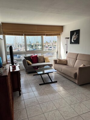 Ein Wohnzimmer mit einer Couch, einem Couchtisch und einer TV-Konsole. Fenster bieten Blick auf Boote und einen Yachthafen.