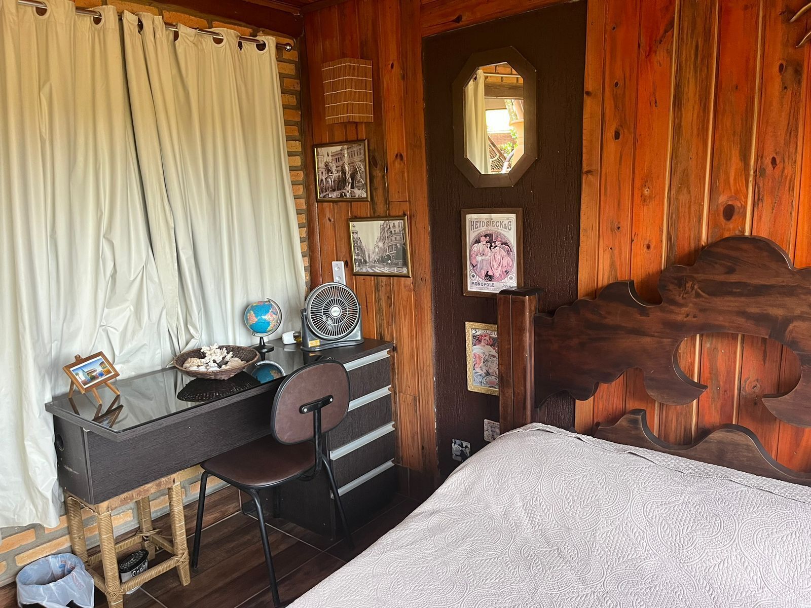 A cozy room with a wooden door, a small desk with a globe and fan, a wicker chair, and a bed with a patterned bedspread.