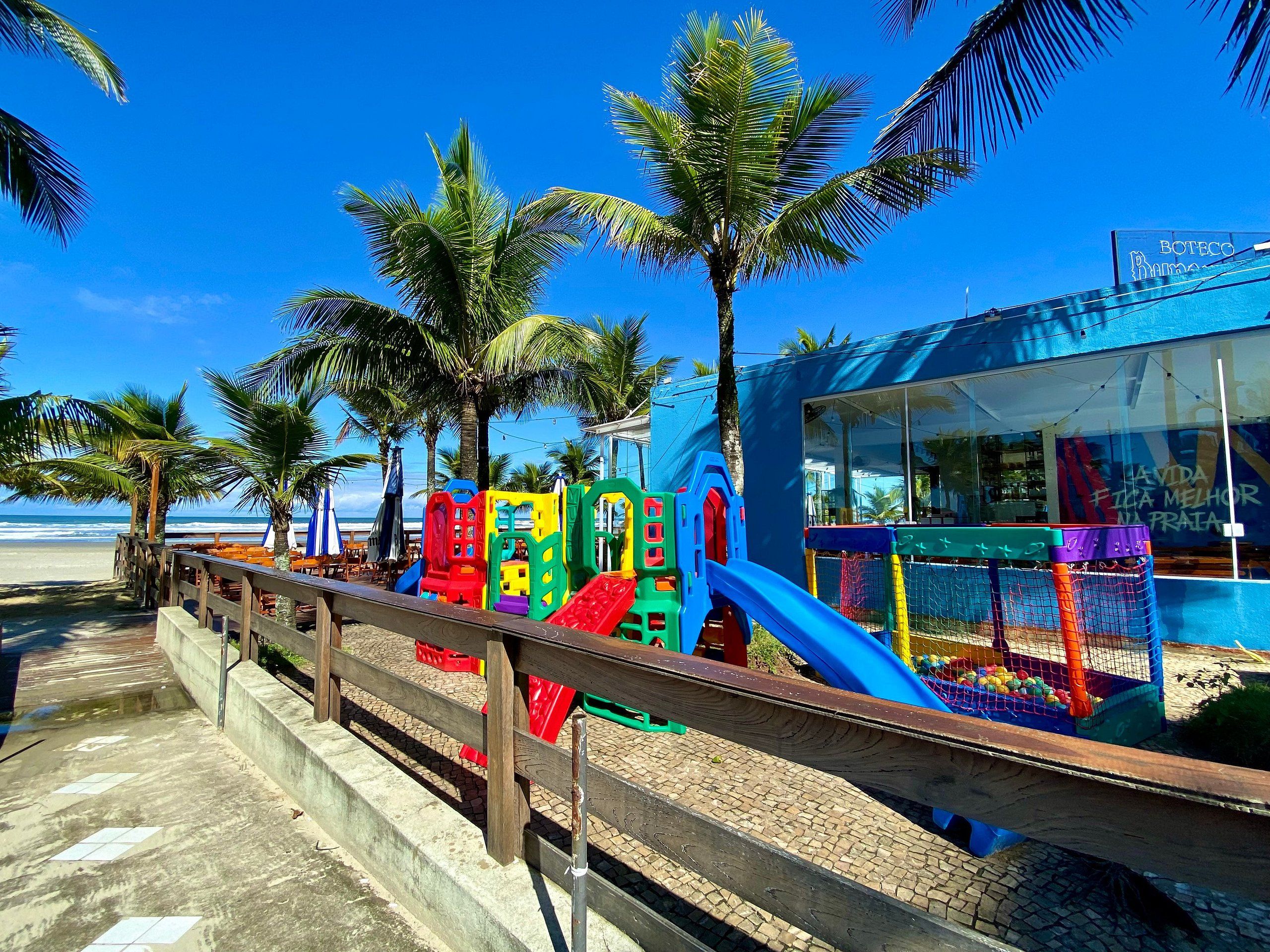 Un parque infantil en la playa con toboganes, estructuras para escalar, palmeras y un edificio con un letrero.
