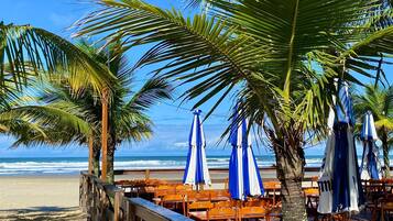 Ein Restaurant direkt am Strand mit Holztischen und blauen Sonnenschirmen mit Blick auf den Ozean.