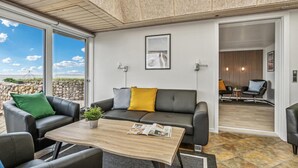 Ein modernes Wohnzimmer mit einer grauen Couch, einem Holztisch und Blick ins Freie durch große Fenster.