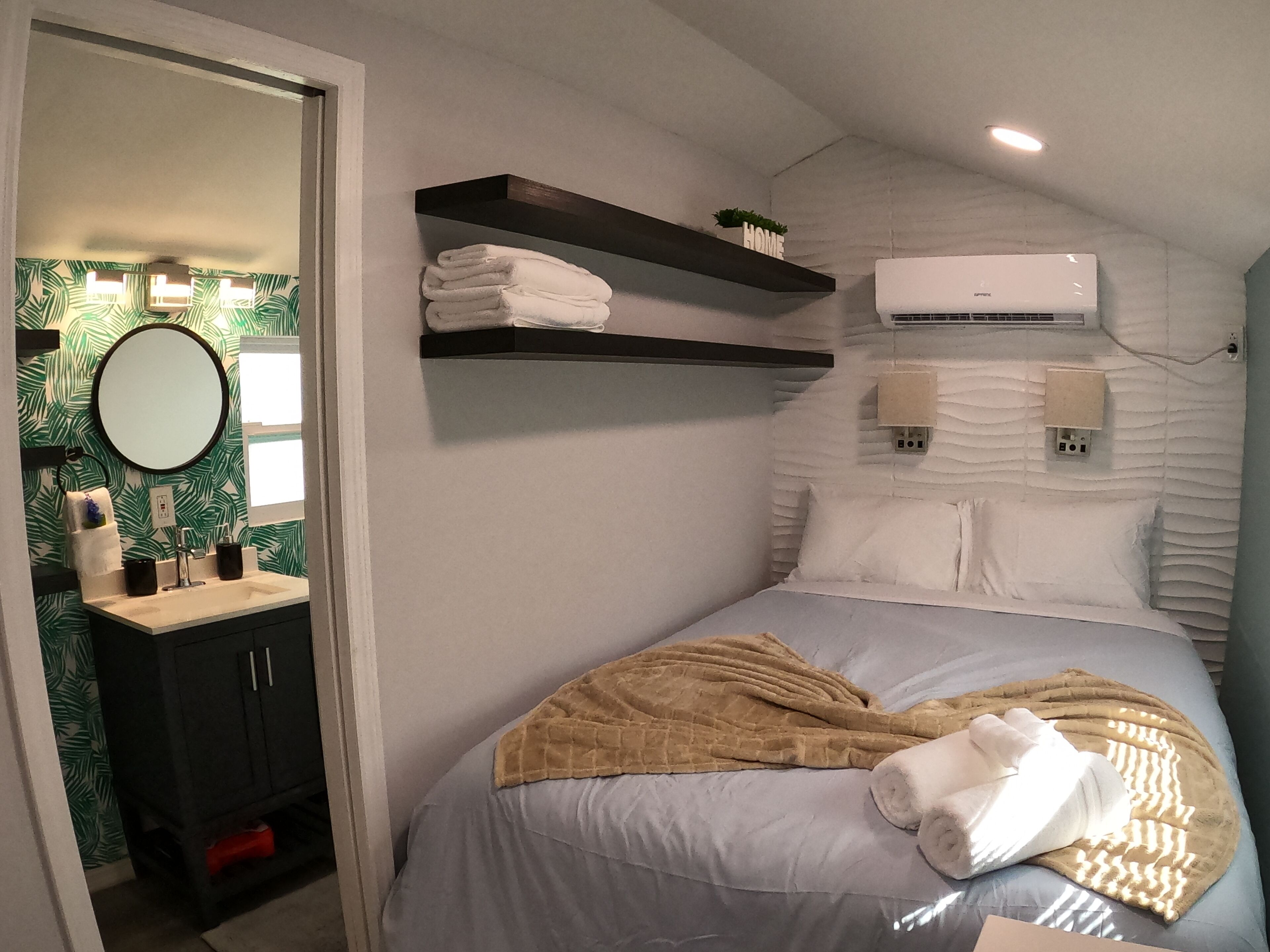 A bedroom with a bed, a bedside table, a mirror, a wall-mounted air conditioner, and a shelf with towels.