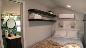 Un dormitorio con una cama, una mesita de noche, un espejo, un aire acondicionado instalado en la pared y un estante con toallas.