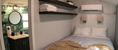 A bedroom with a bed, a bedside table, a mirror, a wall-mounted air conditioner, and a shelf with towels.