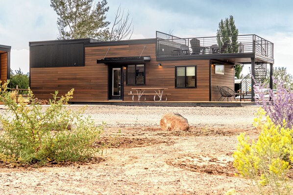 A modern, two-story building with a wooden exterior, a covered patio with seating, and a small garden area with plants and a rock. - Capitol Casita - 2BD+loft+2BA and rooftop deck (Wayne County)