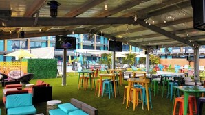 Un espace extérieur aménagé avec des tables et des chaises aux couleurs vives, une pelouse artificielle verdoyante et une structure couverte dotée de poutres apparentes et de luminaires suspendus.