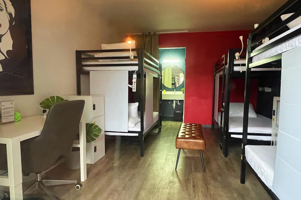 Quarto de hotel pequeno com beliches, escrivaninha, cadeira e um pequeno banco.