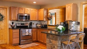 A kitchen with wooden cabinets, a built-in microwave, a stove, a stone island, and bar stools. - ALPINE RIVER ESCAPE - SECLUDED RIVERFRONT CABIN (Sautee Nacoochee)