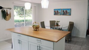 Een keuken met een houten aanrecht, witte kasten, een glazen eettafel met rieten stoelen, en twee schilderijen aan de muur.