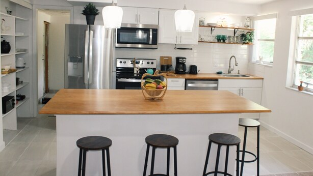 Een moderne keuken met witte kastjes, roestvrijstalen apparatuur, een houten kookeiland met krukken, en hanglampen.
