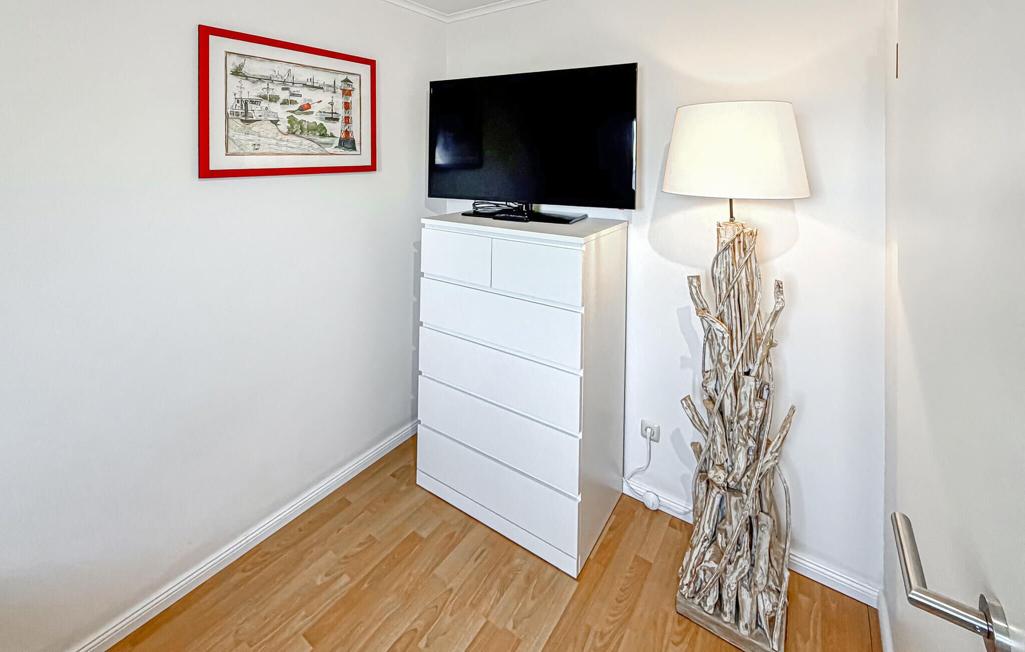 Ein Zimmer mit einem weißen Schminktisch, einem Flachbildfernseher, einer Lampe und einem gerahmten Bild an der Wand.