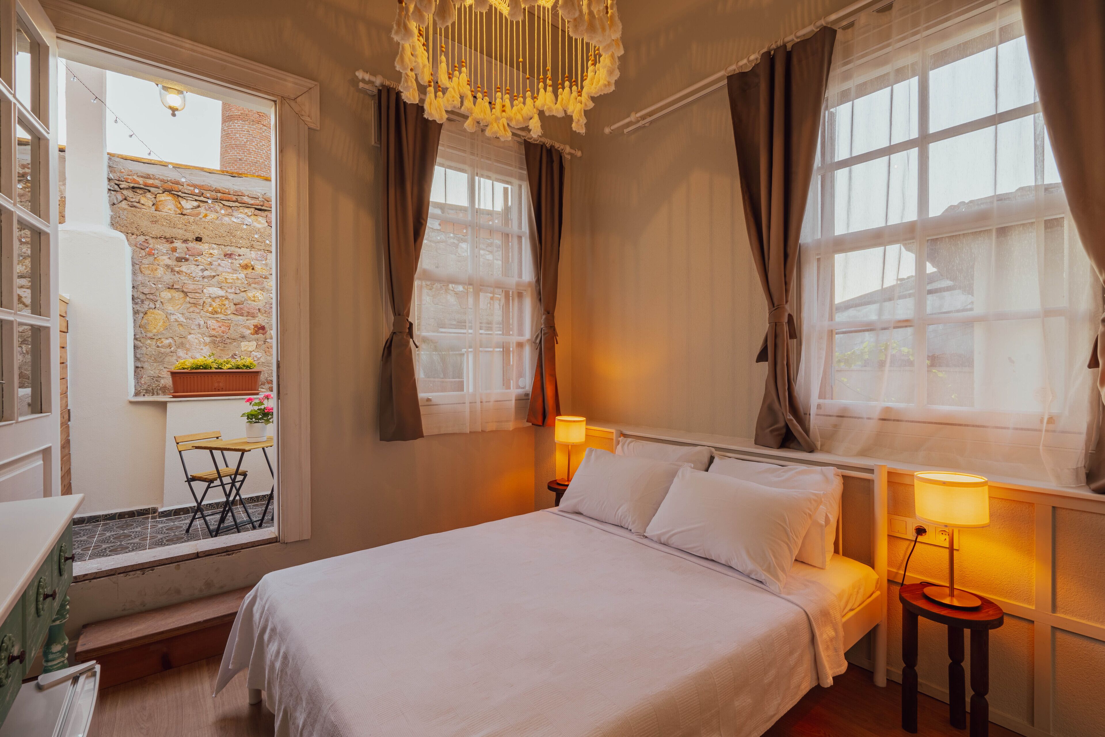 Un dormitorio con cama, lámparas de noche, una ventana con cortinas, una mesita con una silla y vistas a un muro de piedra.
