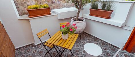 Un pequeño espacio al aire libre con una mesa de madera, dos sillas, una planta en maceta y un taburete blanco.