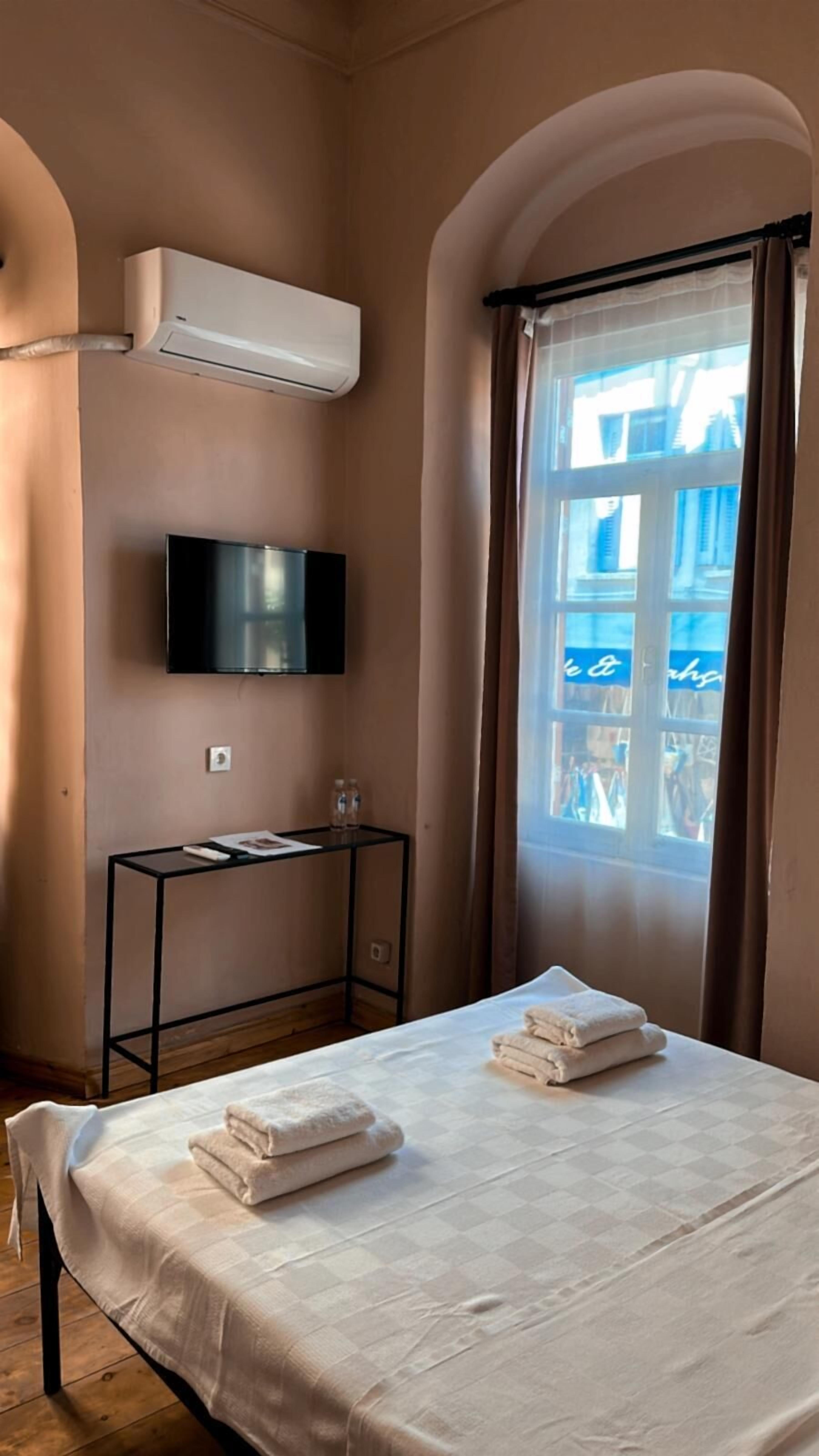 A hotel room with a bed, a flat-screen TV mounted on the wall, a small table with a magazine, and a window with curtains.