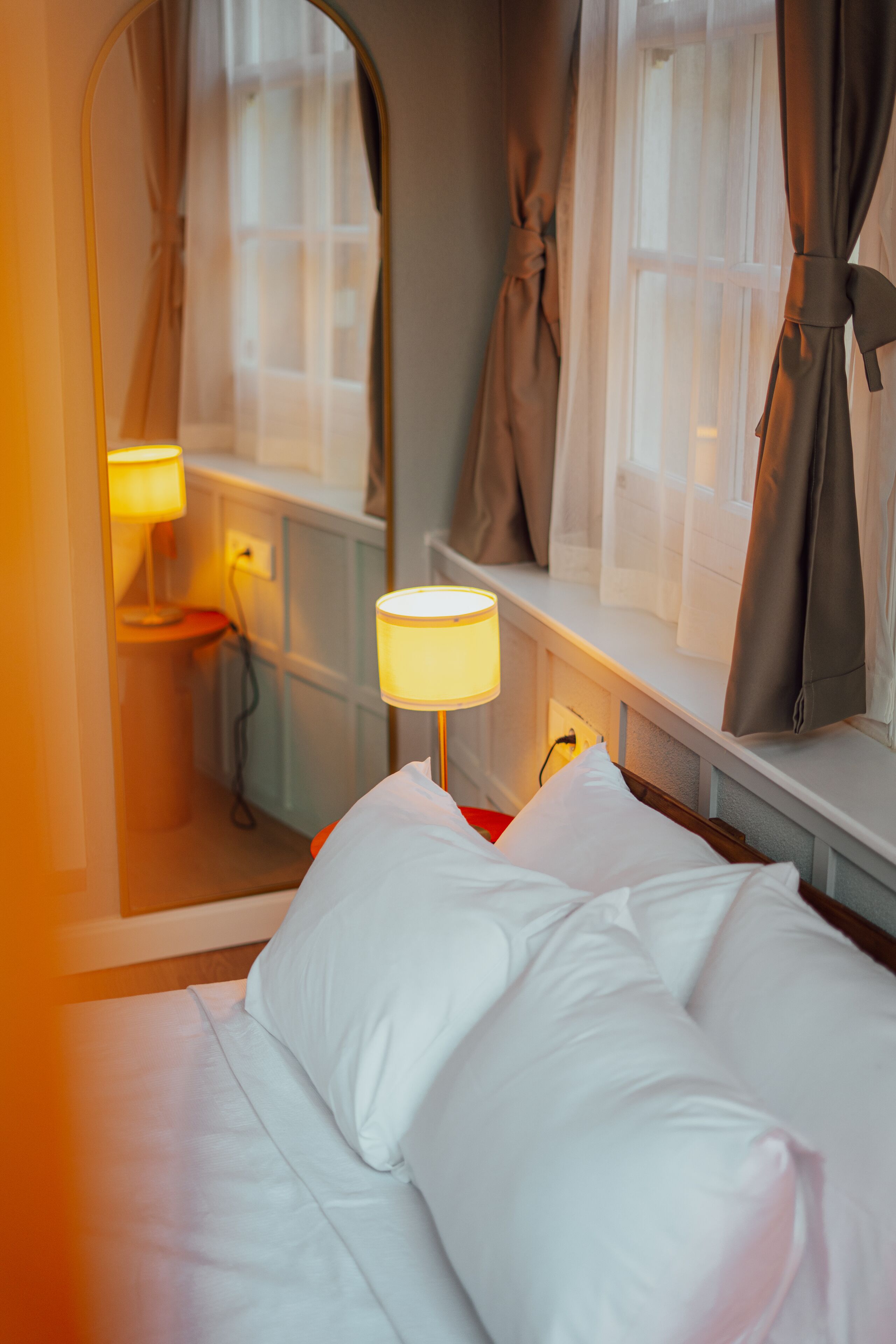 A hotel room with a bed, a bedside table with a lamp, a window with curtains, and a mirror on the wall.