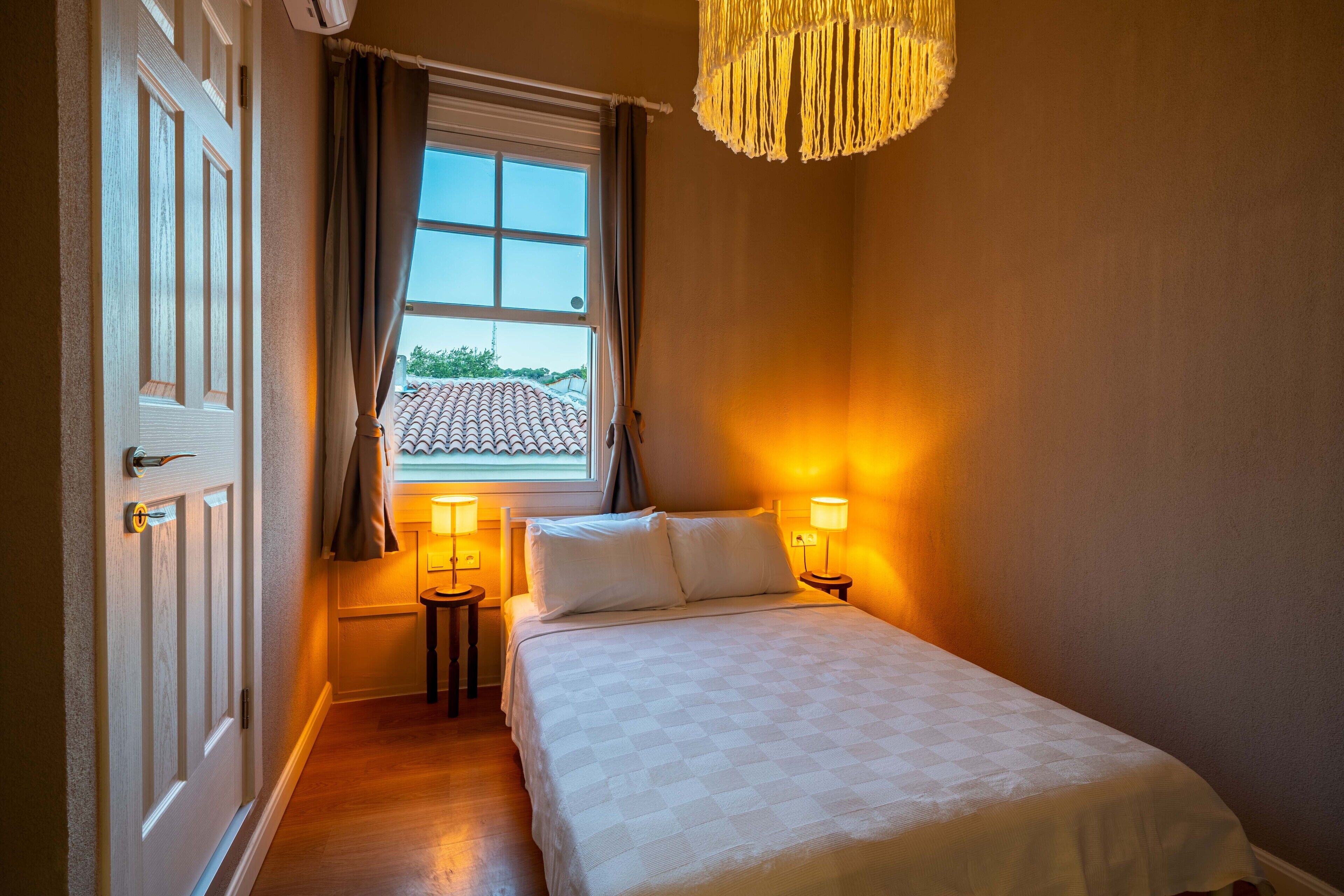 A bedroom with a bed, two bedside lamps, a window with curtains, and a hanging light fixture.