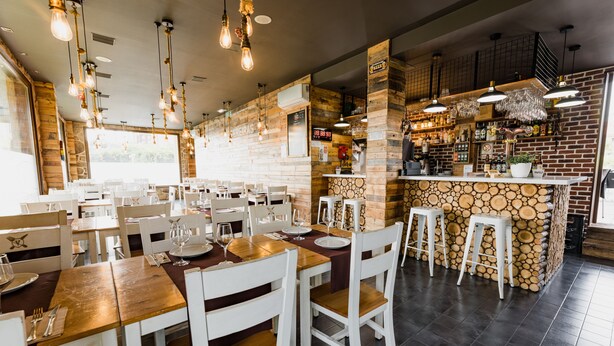 A restaurant with wooden tables and white chairs, a bar area with stools, and a stone wall.
