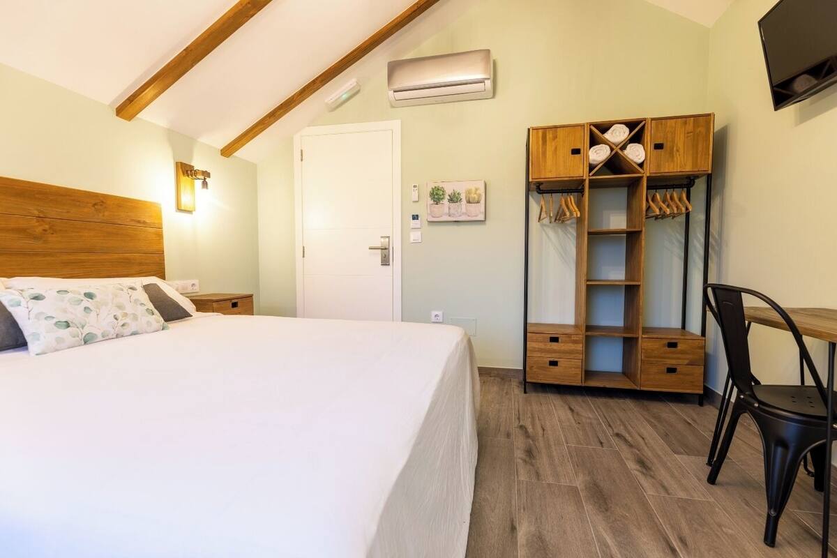 A bedroom with a bed, a wooden wardrobe, a desk with a chair, and an air conditioning unit.