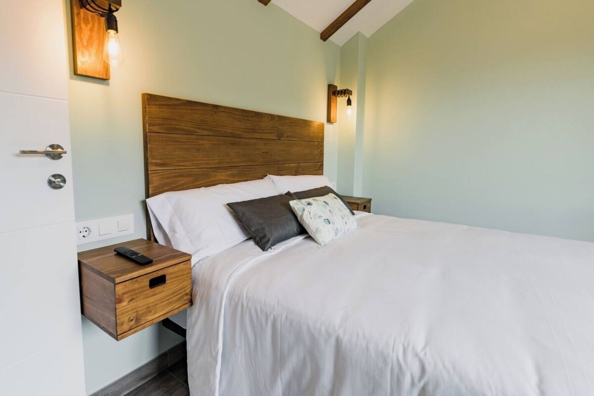 A neatly made bed with white linens and two pillows, a wooden nightstand with a remote control, and a wall-mounted light fixture.