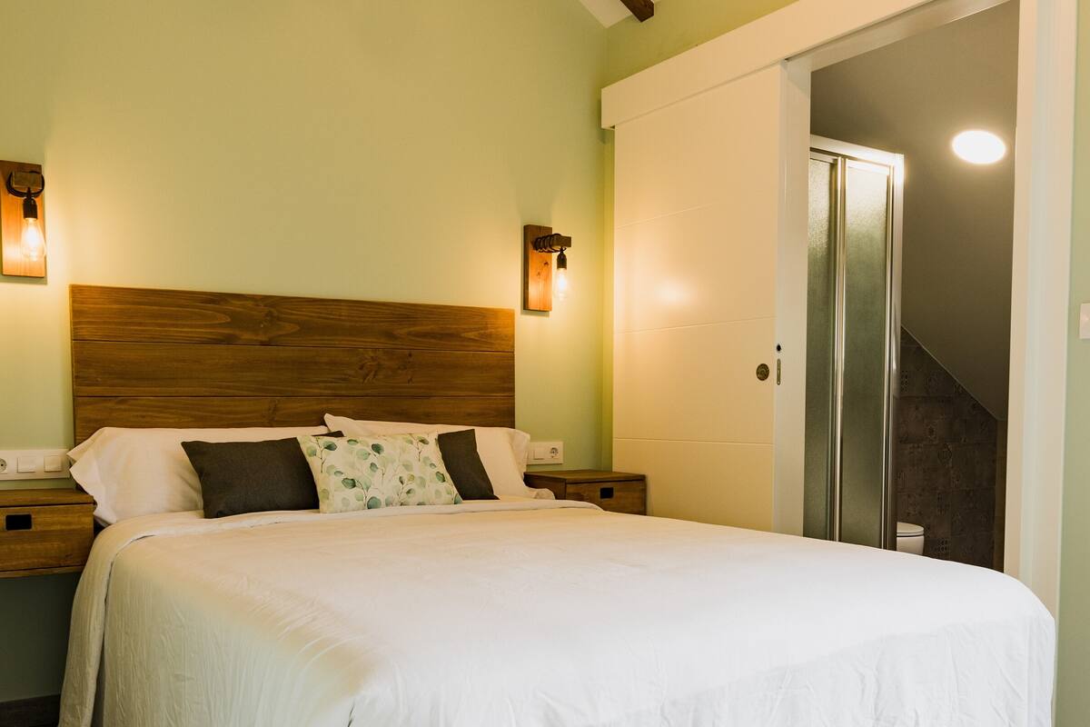 A neatly made bed with white bedding and a wooden headboard, two bedside tables with lamps, and a door leading to a bathroom.