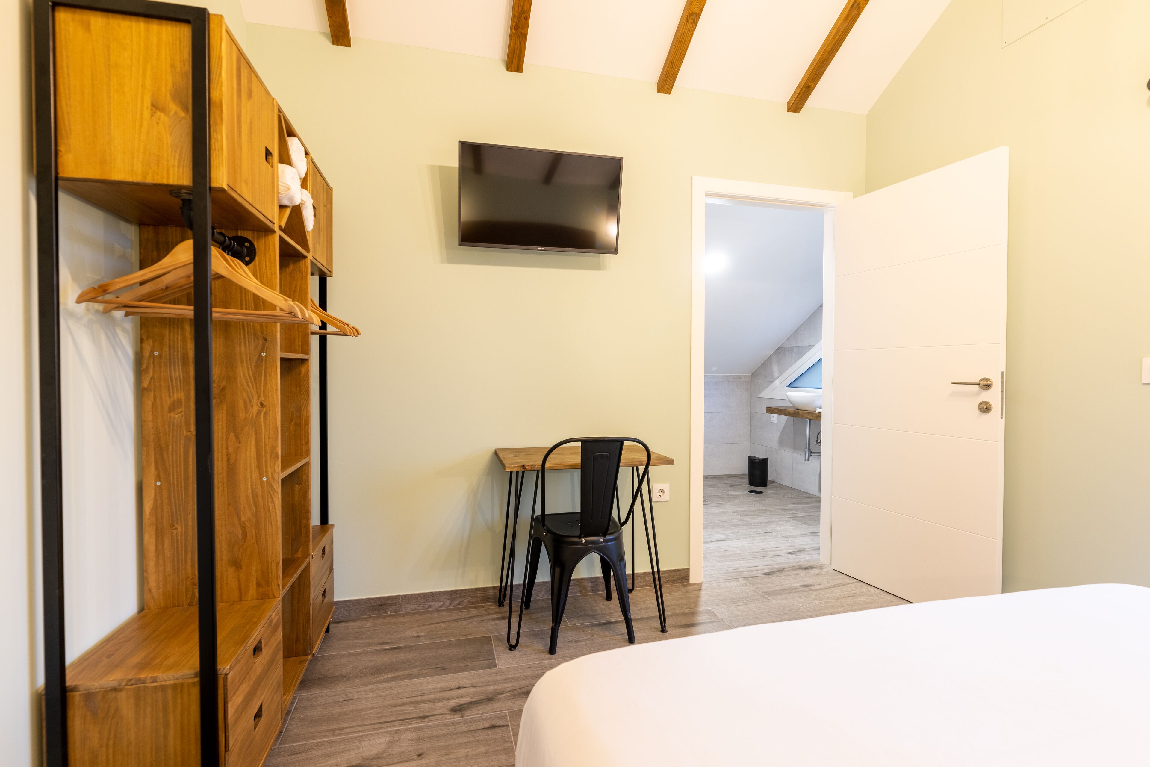A bedroom with a bed, wooden wardrobe, a desk with a chair, and a TV on the wall.