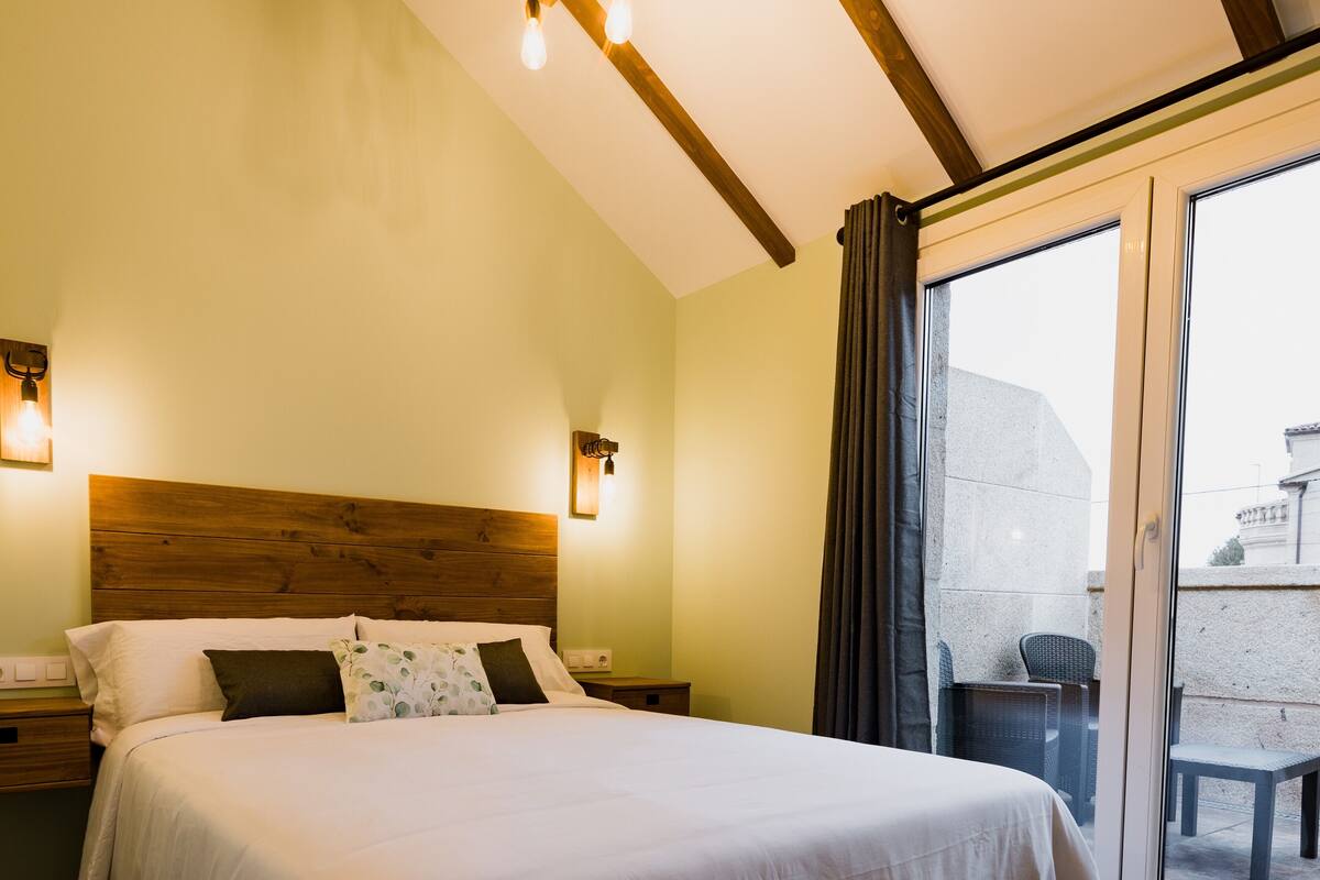 A hotel room with a wooden headboard, a large bed with white bedding, two bedside lamps, and a view of the outside through a glass door.