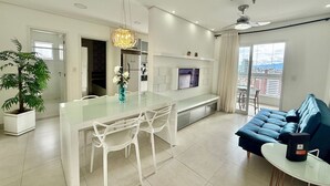 A modern living room with a glass dining table, white chairs, a blue sofa, a TV, and a balcony with a city view. - Flat Unitá Homestay (Praia Grande)