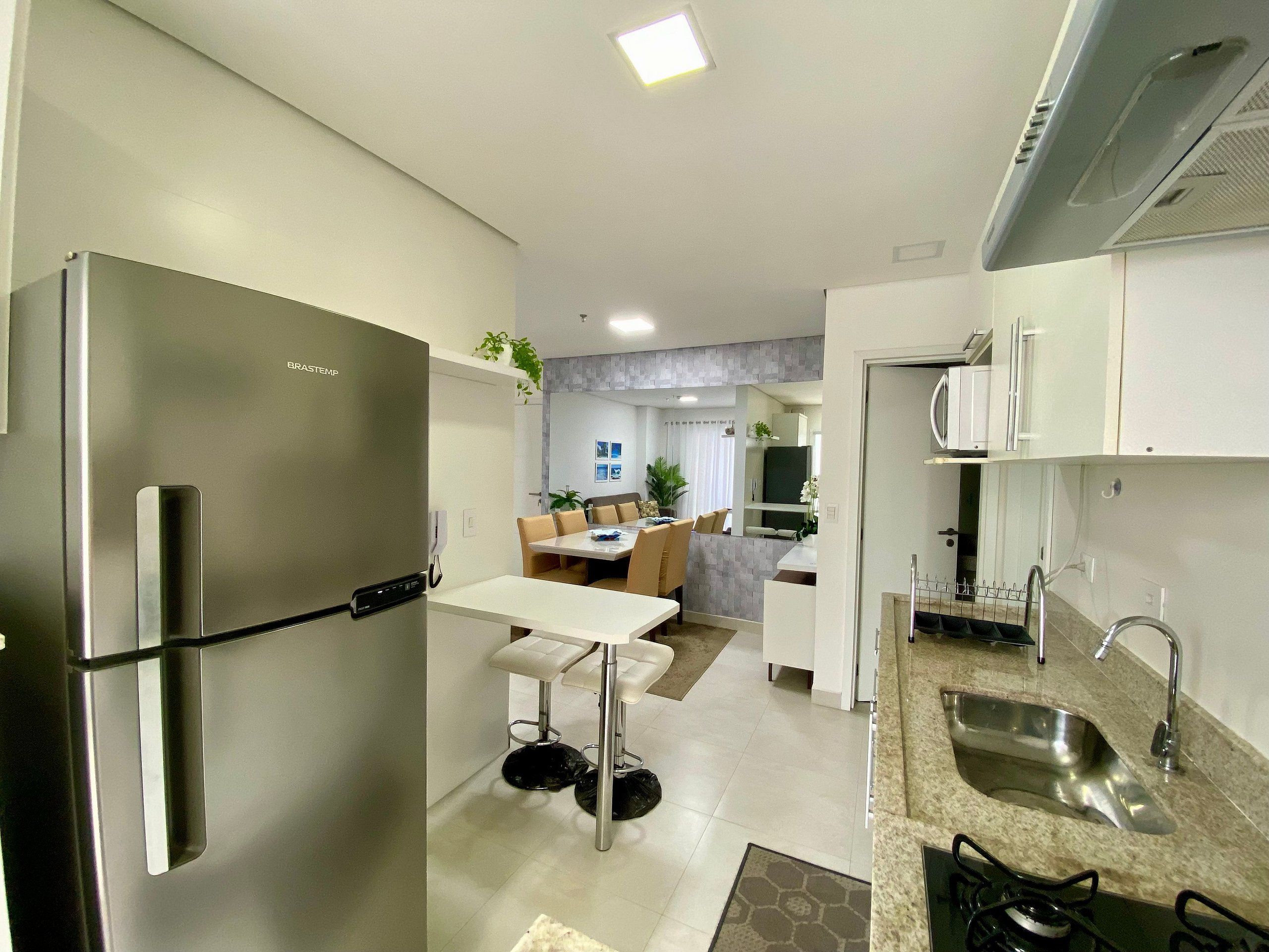 Uma cozinha moderna com geladeira de aço inoxidável, uma área de jantar com mesa e cadeiras brancas, e um espaço de estar visível com sofá e televisão.