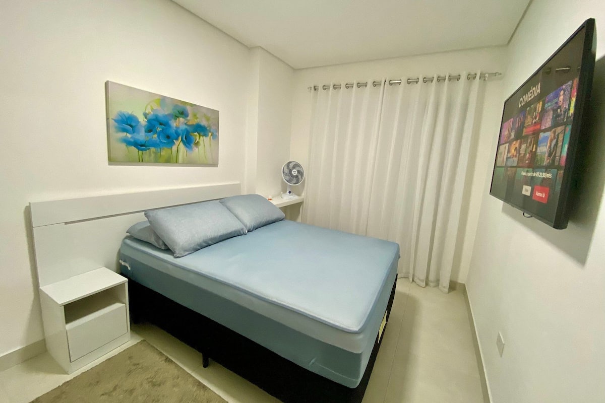 A bedroom with a bed, a nightstand, a wall-mounted TV, and a painting.