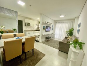 A modern living room with a sofa, a dining area with a table and chairs, a kitchen with cabinets and a TV, and a large window with curtains. - Flat Unitá Homestay (Praia Grande)