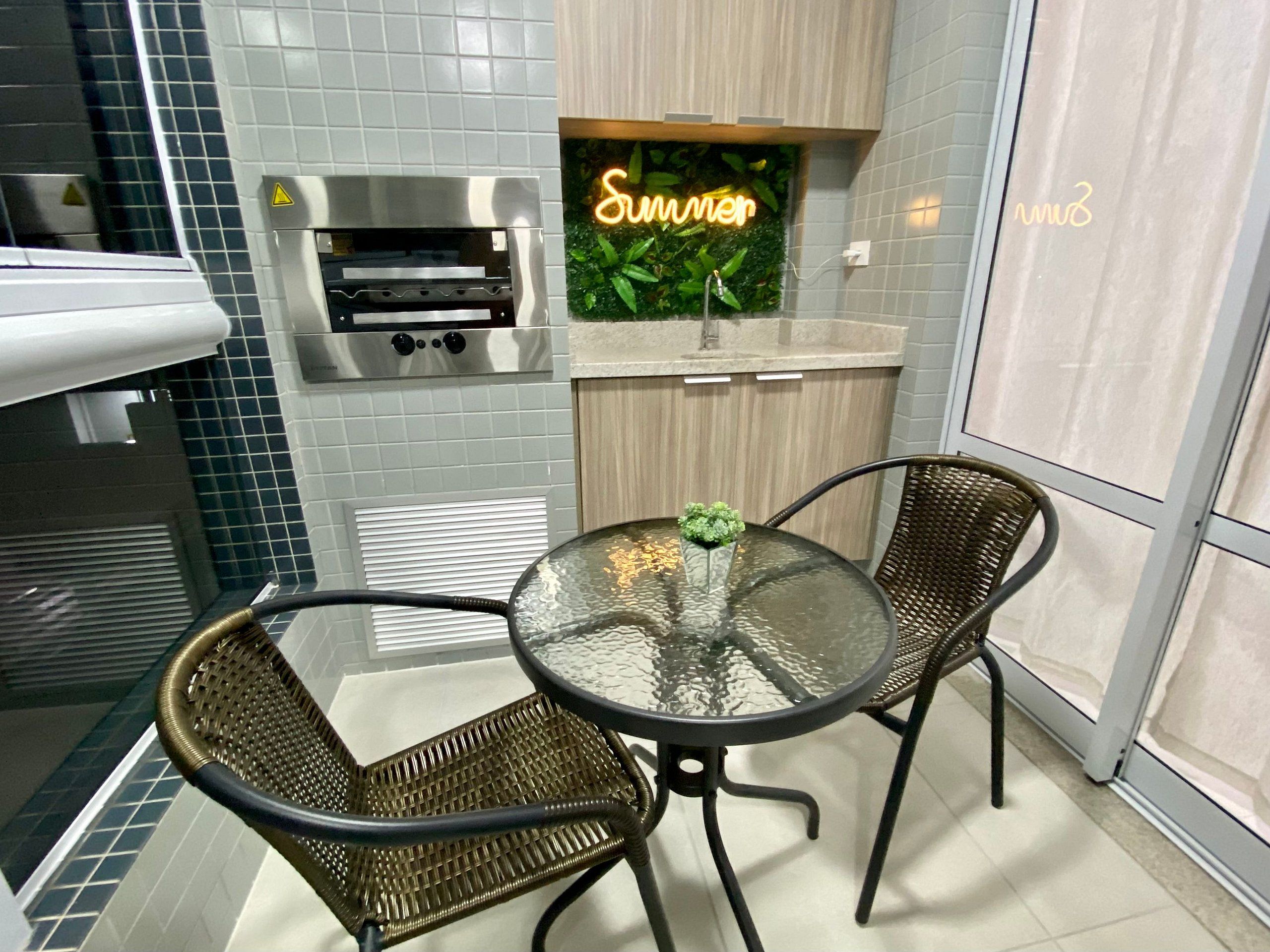 A small kitchen with a built-in oven, a glass table with chairs, and a neon sign reading 'Summer.'