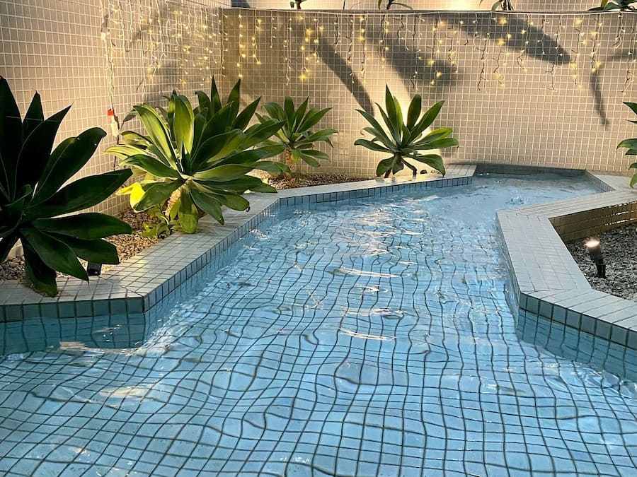 A swimming pool with tiled flooring, surrounded by plants and a tiled wall.