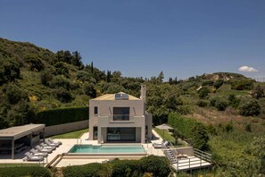 A modern two-story house with a swimming pool, surrounded by lush greenery and hills.