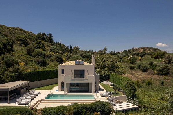 Ein modernes, zweistöckiges Haus mit Swimmingpool, umgeben von üppiger Vegetation und Hügeln.