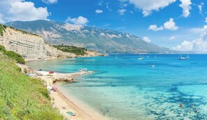 Ein Küstenstrand mit klarem türkisblauem Wasser, einer felsigen Küstenlinie und einer gebirgigen Kulisse.
