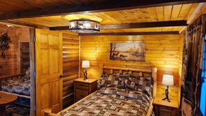 A cozy log cabin bedroom with a bed, two bedside tables, a nightstand, a wall clock, and a painting of deer. - Waterfront Cabin - Hot Tub - Free Bikes, Kyaks, Canoes, Paddle, & Fishing Boats! (Waterville)