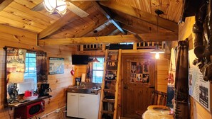 A rustic cabin interior with wooden walls, a kitchenette, a mounted television, and various decorations. - Waterfront Cabin - Hot Tub - Free Bikes, Kyaks, Canoes, Paddle, & Fishing Boats! (Waterville)
