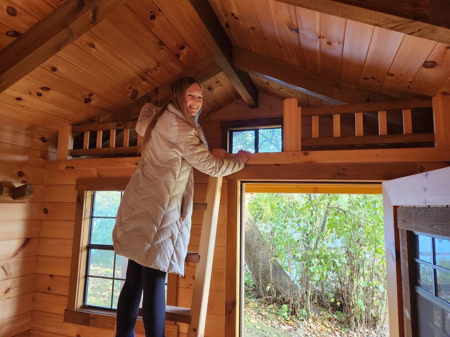 A person standing on a wooden beam inside a cabin with a view of trees through a window.