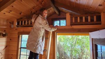 A person standing on a wooden beam inside a cabin with a view of trees through a window.