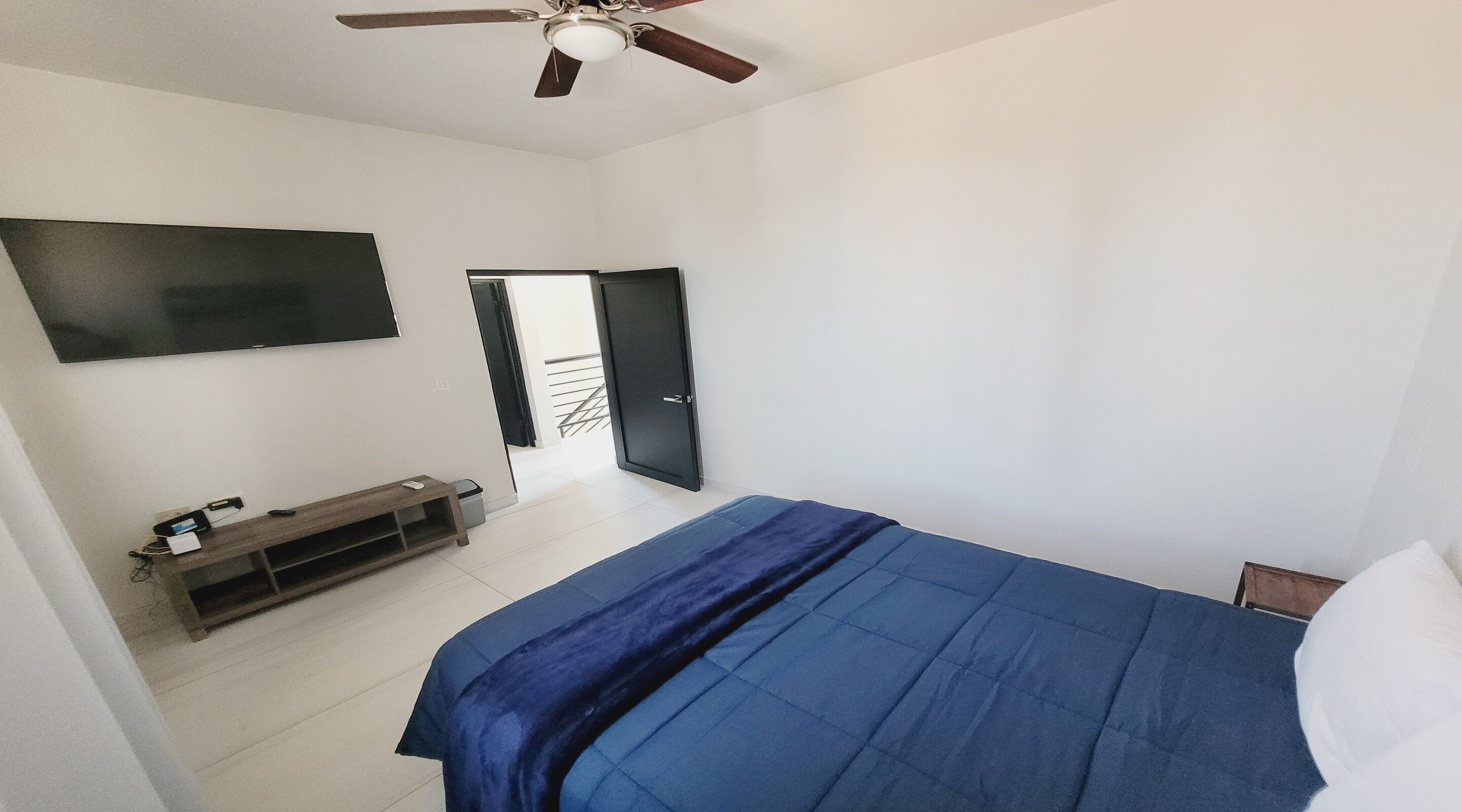 A bedroom with a bed, a TV mounted on the wall, a ceiling fan, and a door leading to a balcony.
