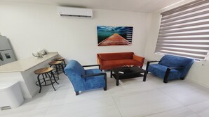 A compact living space with a kitchenette, a dining area with stools, a sofa, a coffee table, and a large window with blinds. - Casamar 4 by Kivoya (Puerto Peñasco)
