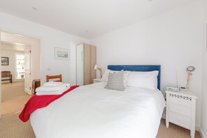 Habitación de hotel con una cama grande, una mesita de noche con una lámpara y otro buró con un dispositivo digital, y una puerta que da a un baño.