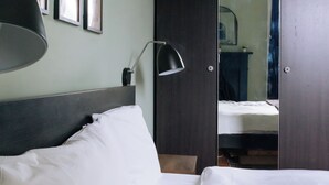A neatly made bed with white linens, a dark headboard, and a wall-mounted lamp. There are framed artworks on the wall and a mirror reflecting a bed and a dresser. - Stylish 1BD Flat - Abbeyhill (Edinburgh)