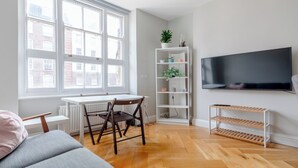 Moderni olohuone, jossa on valkoinen patteri, ruokapöytä tuoleineen, harmaa sohva, seinälle asennettu televisio ja puulattia.