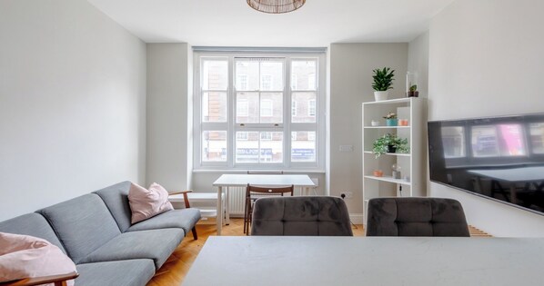 Un salon moderne avec un canapé gris, une table à manger et des chaises. Une grande fenêtre offre une vue sur l’extérieur.