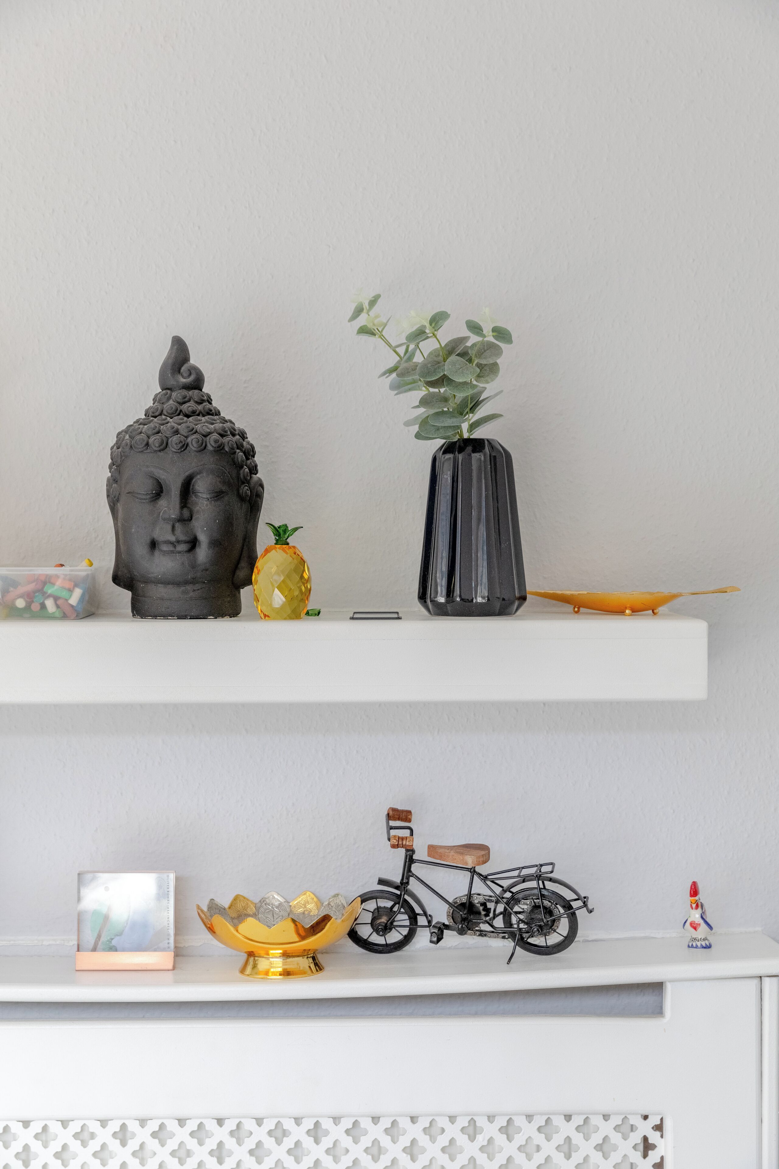 Una estanteria amb una cap Buddha, una pinya, un vas amb fulles verdes, un bol petit, una bicicleta i una figura.