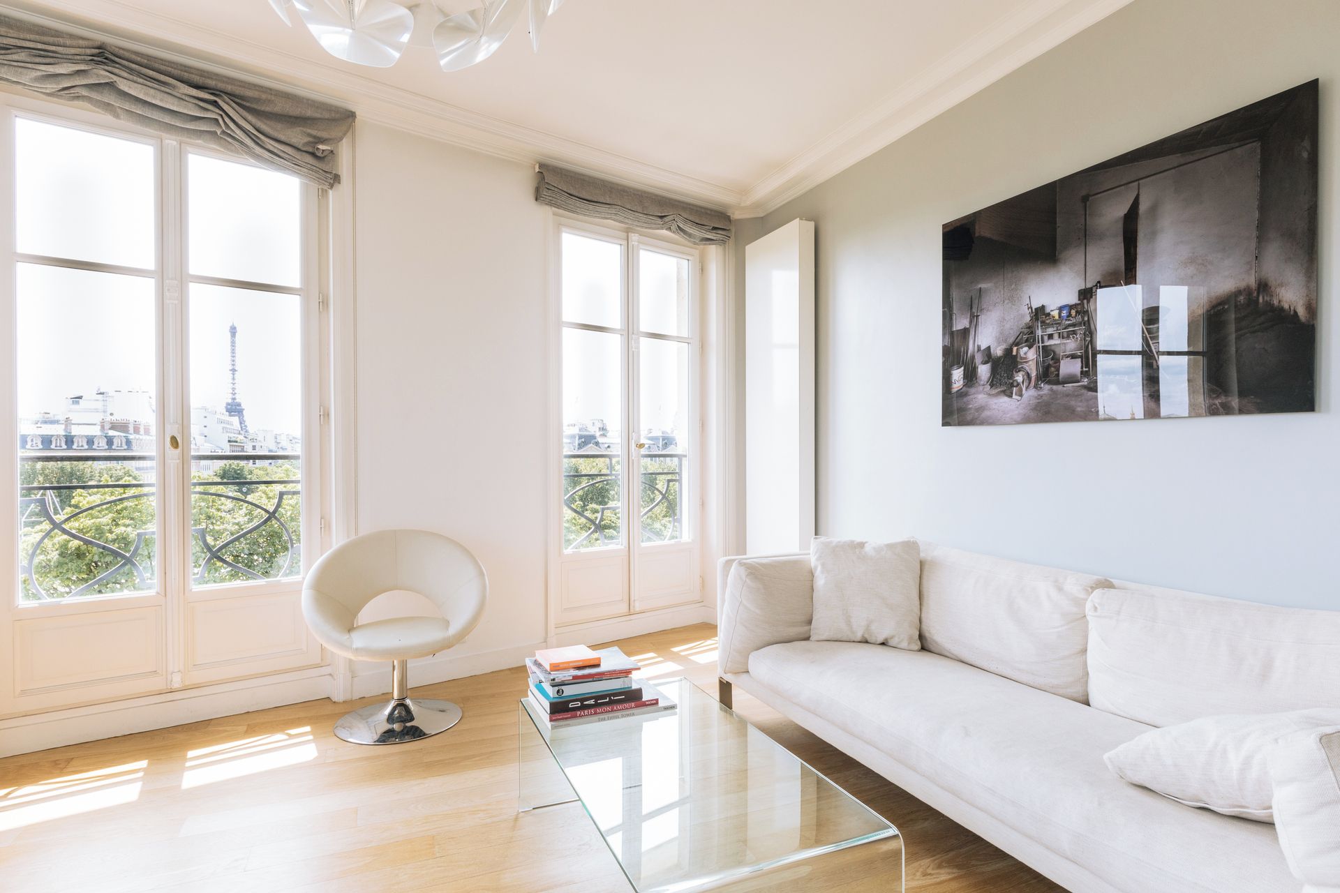 A modern living room with a white sofa, a glass coffee table, and a chair. There are large windows with a view of a cityscape.